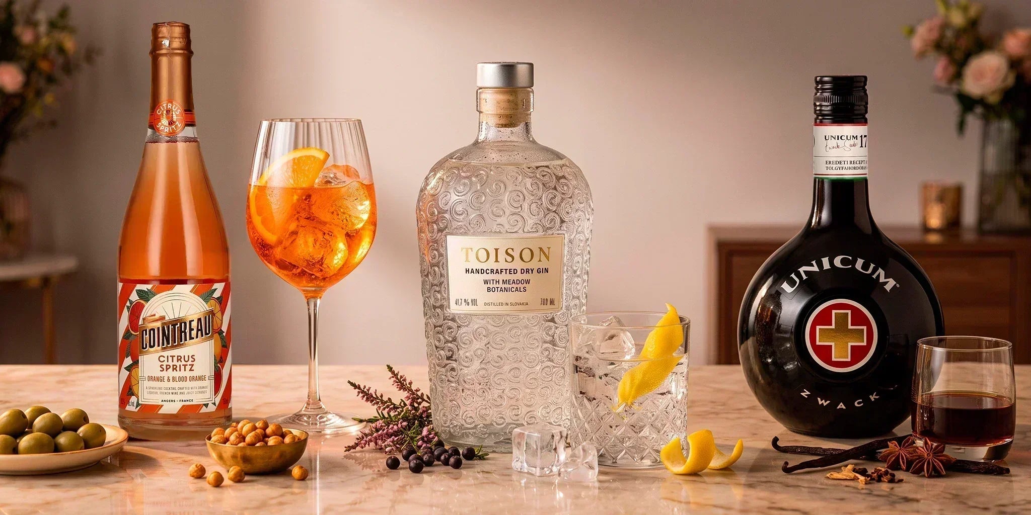 Three-bottle hosting formula with Cointreau Citrus Spritz, Toison Handcrafted Dry Gin and Zwack Unicum, styled with cocktails, citrus, olives, herbs and spices on a marble table.