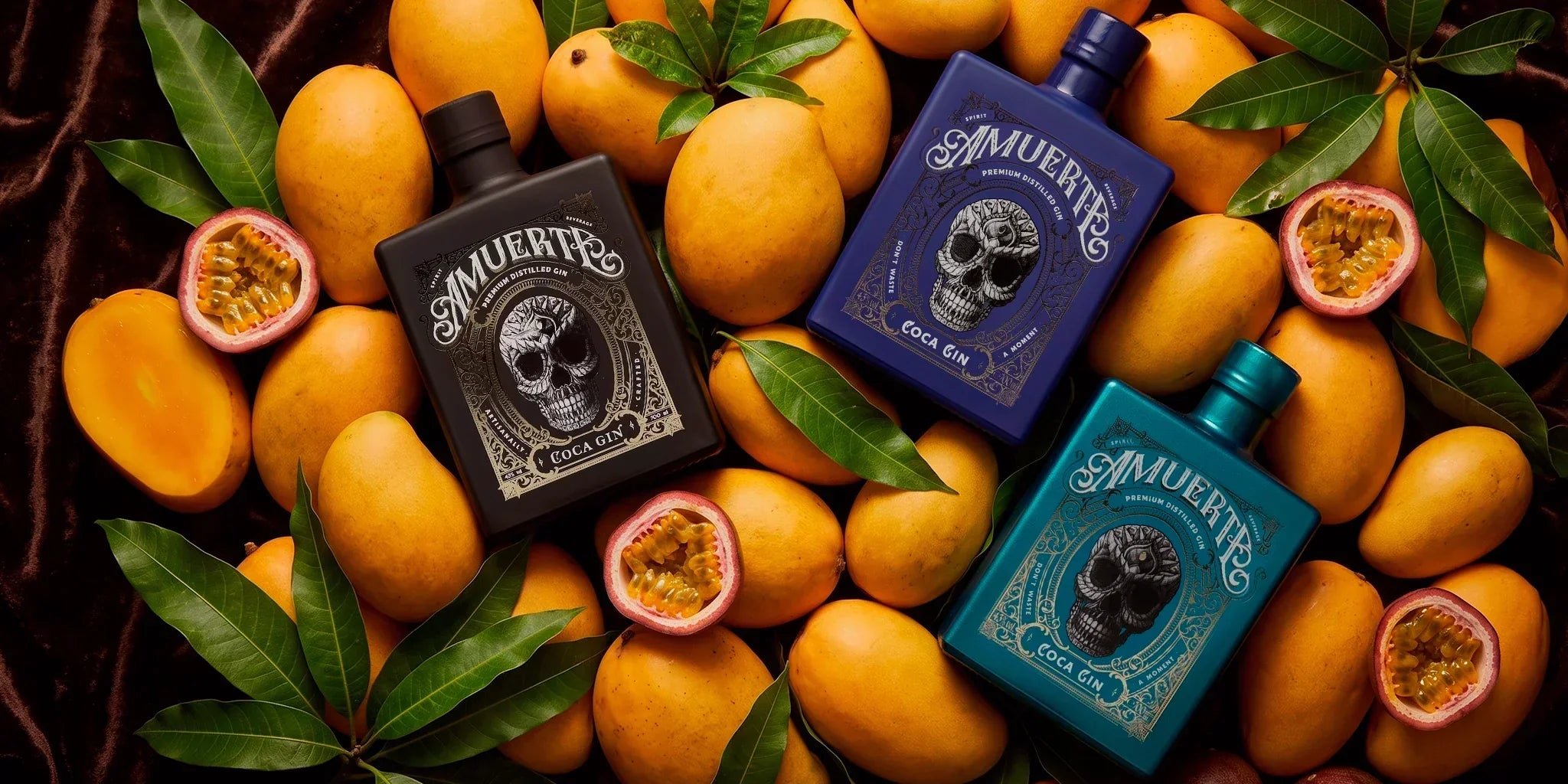 Amuerte Coca Gin collectible bottles in black, blue and turquoise editions styled on tropical mangoes and passion fruit with green leaves, creating a bold premium gin flat lay for rare bottle collectors.