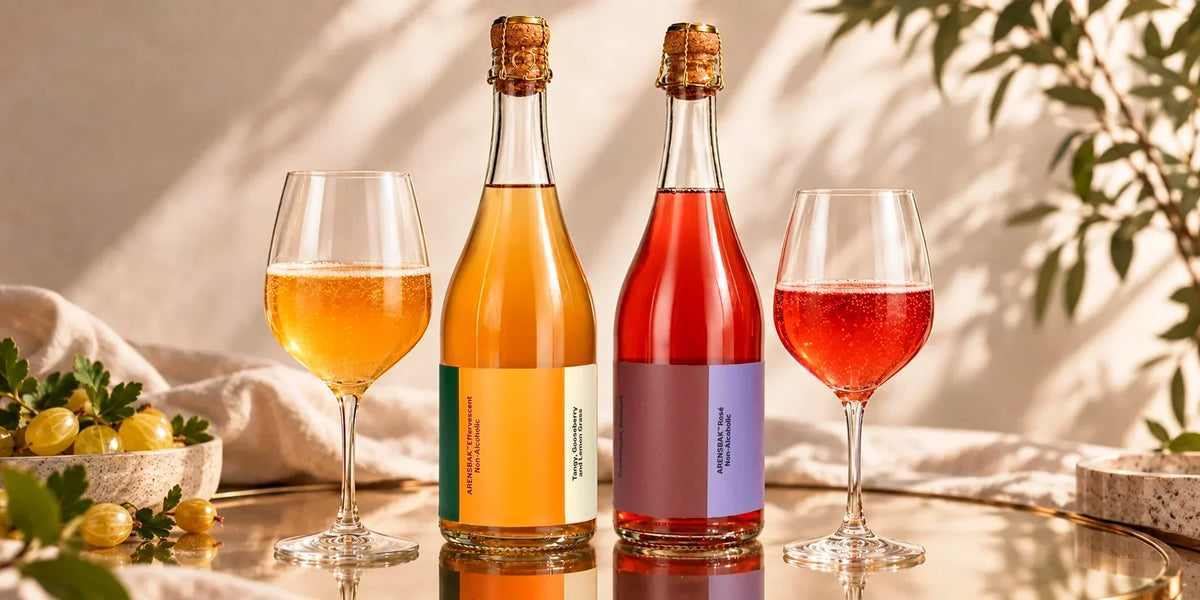 ARENSBAK non-alcoholic sparkling wine bottles, including Effervescent and Rosé, served with matching glasses in a bright elegant setting with fresh gooseberries and soft natural light.