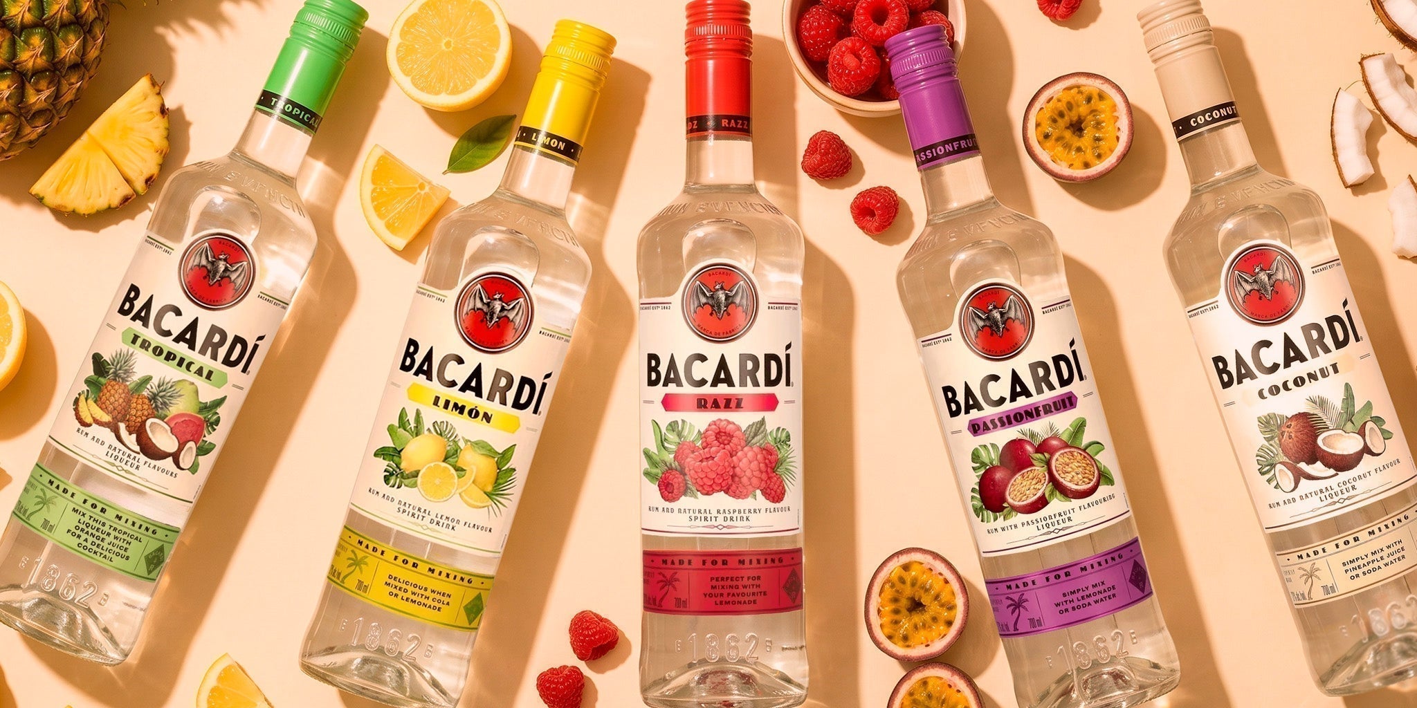 Bacardi flavored rum bottles including Tropical, Limón, Razz, Passionfruit and Coconut with fresh pineapple, lemon, raspberries, passion fruit and coconut on a sunny summer background.