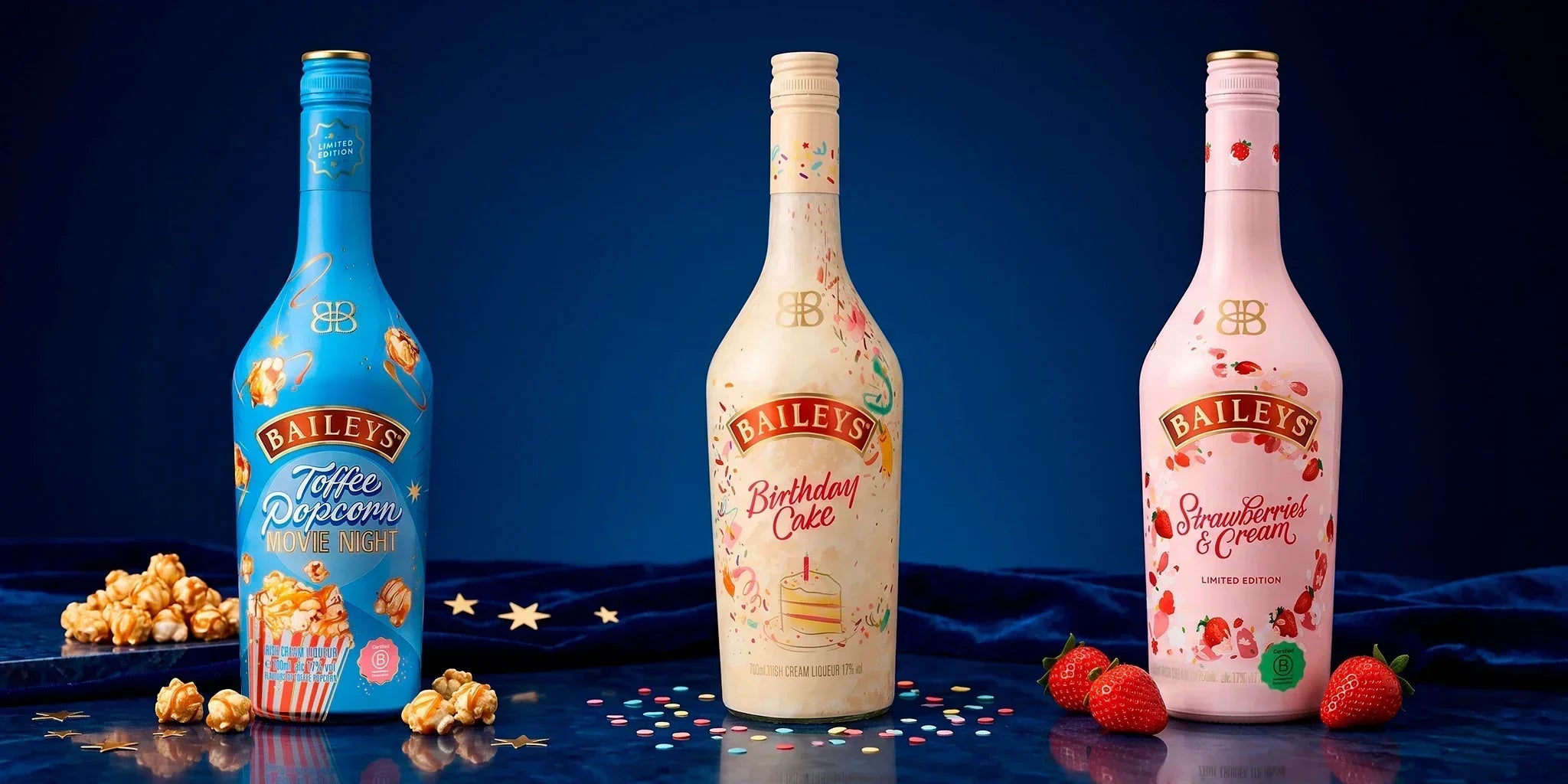 Baileys dessert liqueurs selection with Toffee Popcorn Movie Night, Birthday Cake and Strawberries & Cream bottles styled with popcorn, confetti and strawberries on a dark blue background.