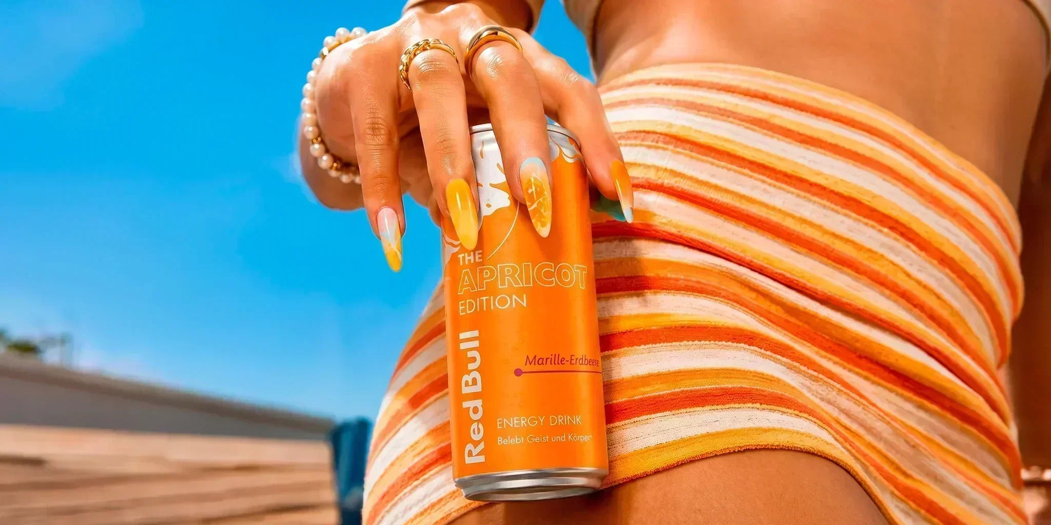 Red Bull The Apricot Edition energy drink can held against a bright blue summer sky, with orange striped outfit and matching apricot-colored manicure for a bold sunny lifestyle image.
